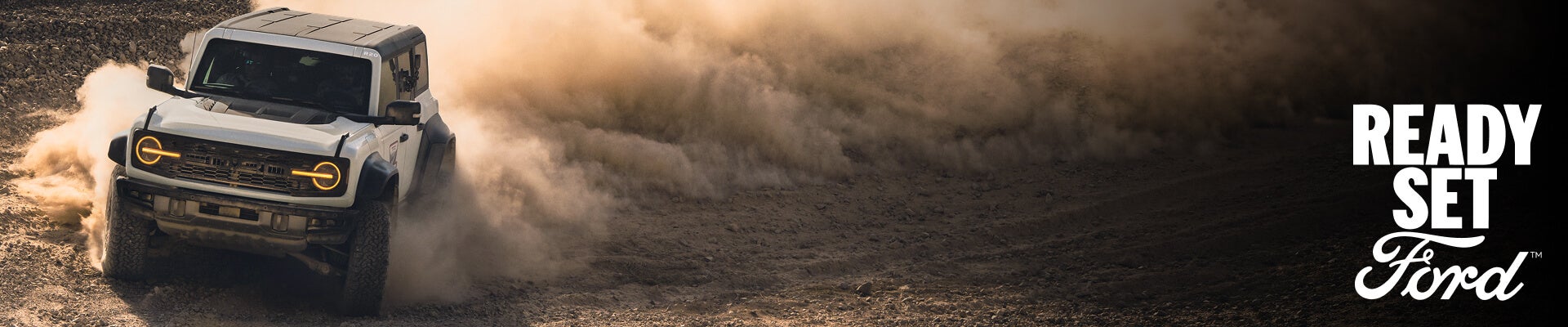Front three-quarter view of a 2027 Ford Bronco Raptor in Oxford White kicking up dust on an off-road desert trail.